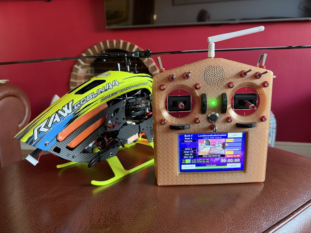 A yellow and black Goblin RAW 420 helicopter on a table beside a gold-coloured LockDownRadioControl transmitter with two sticks, eight switches and its Version 1 screen lit
